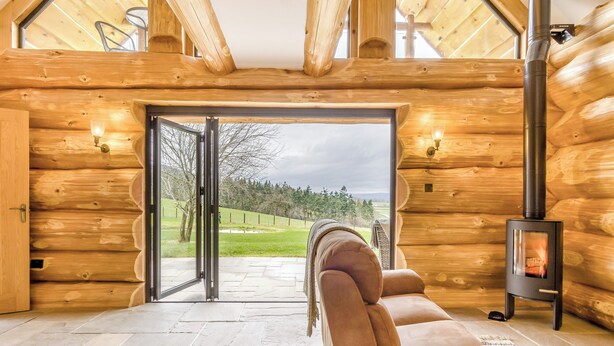 L’intérieur d’une cabane en rondins, avec un sol en pierre, un canapé, une cheminée et une porte vitrée donnant sur l’extérieur.