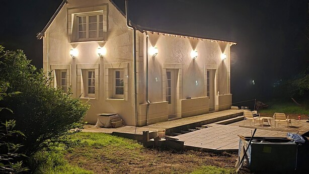 Une maison à deux étages, dotée d’un toit en pente, de nombreuses fenêtres et d’une terrasse dallée aménagée avec du mobilier d’extérieur.