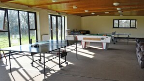 Una habitación con mesa de ping pong, mesa de billar y mesa de futbolín. Amplias ventanas permiten la entrada de luz natural y vistas al exterior.