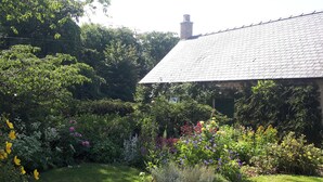 Een klassiek stenen huis met schuin dak, omgeven door een weelderige tuin met verschillende bloeiende planten en een verzorgd gazon.