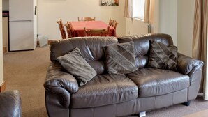 A living room with a brown leather sofa, a dining table with chairs, a refrigerator, and a painting on the wall.