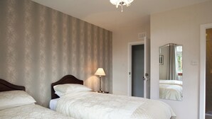 Une chambre d’hôtel avec deux lits, une table de chevet avec une lampe, une porte donnant sur une salle de bain et un miroir qui reflète la pièce.