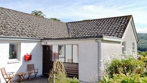 Un bâtiment blanc avec un toit recouvert de tuiles, une porte d’entrée et un petit jardin agrémenté de plantes et d’un banc.