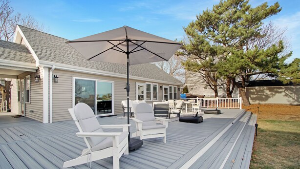 Une terrasse avec deux chaises blanches et un grand parasol, une maison avec un porche abrité, et un arbre en arrière-plan.