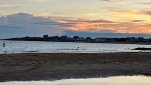 Une plage au coucher du soleil, avec une eau calme et une côte lointaine parsemée de bâtiments.