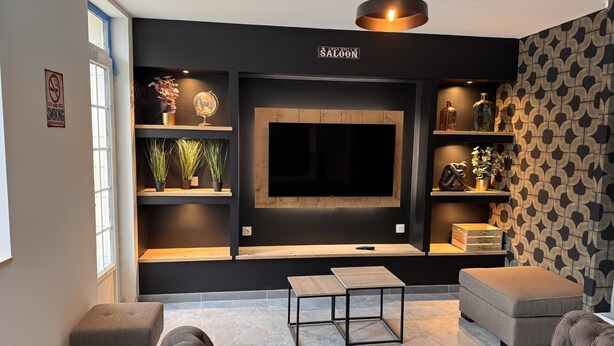 Ein modernes Wohnzimmer mit einem großen Flachbildfernseher an einer dunklen Wand, Holzregalen mit Dekoration und einer gemusterten Tapete auf der rechten Seite.