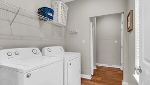 A laundry room with two white front-loading washers and dryers, a white metal shelf with baskets, and a wooden floor. - Blast at the Beach - Golf Cart Available to Rent! Pet Friendly! (Destin)
