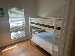 A bunk bed with a white frame and a lower bunk with a quilted mattress. There is a window with blinds to the left, and a wall-mounted electrical outlet is visible. - Blast at the Beach - Golf Cart Available to Rent! Pet Friendly! (Destin)