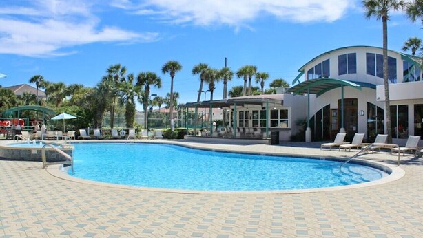 A swimming pool with lounge chairs, palm trees, and a building with large windows. - Blast at the Beach - Golf Cart Available to Rent! Pet Friendly! (Destin)