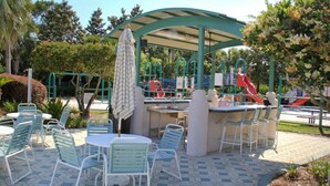 An outdoor patio area with green metal chairs and tables, a covered seating area, and playground equipment in the background. - Blast at the Beach - Golf Cart Available to Rent! Pet Friendly! (Destin)