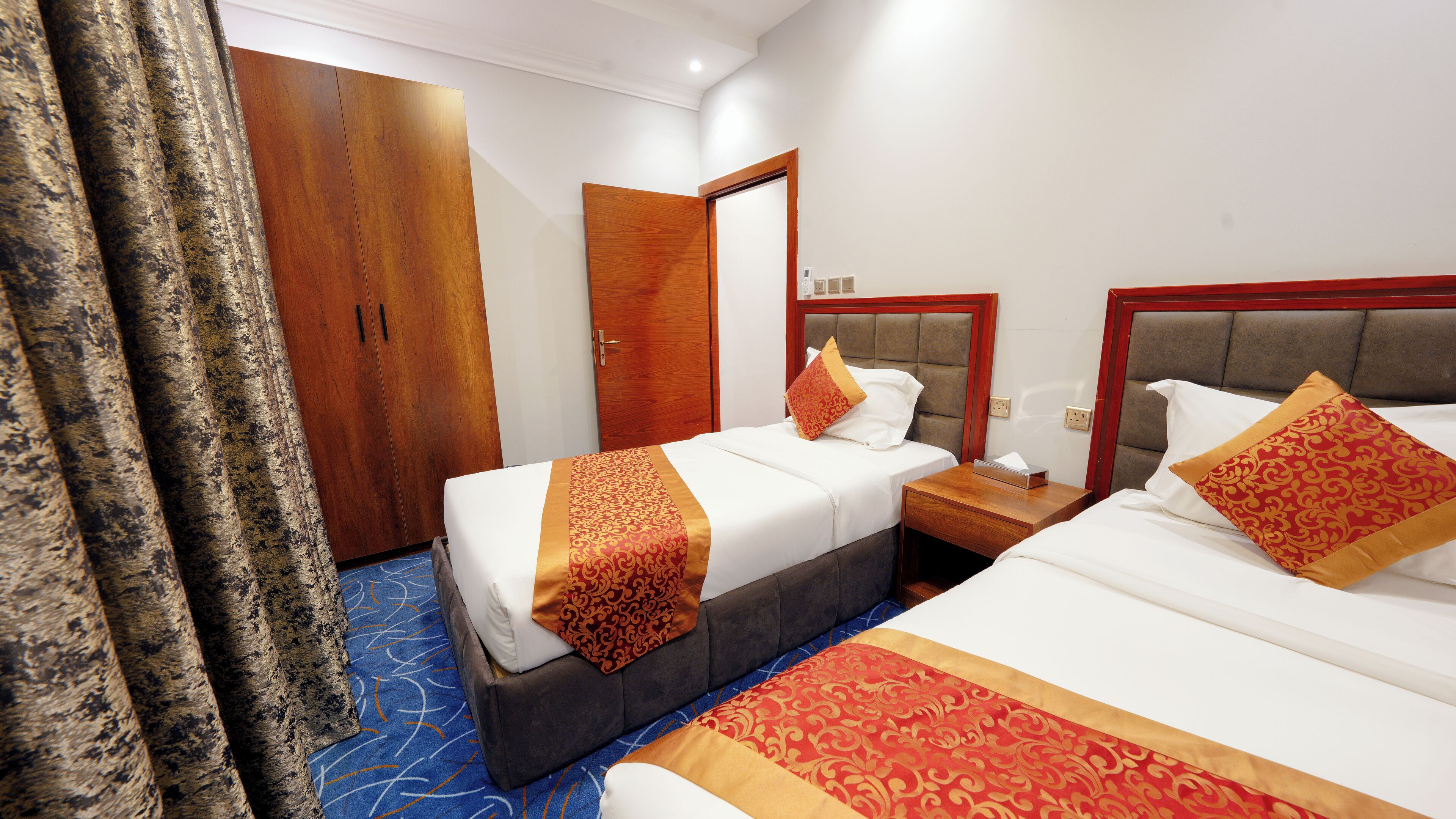 A hotel room with two beds, a wooden wardrobe, a mirror, and a bedside table with a lamp.