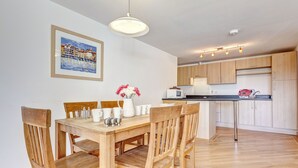 Un coin repas avec une table et des chaises en bois, un pichet blanc orné de fleurs et, au fond, une cuisine.