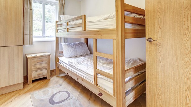 Un lit superposé avec des montants en bois et une literie blanche, une armoire en bois, une petite table de chevet et une fenêtre avec des rideaux.