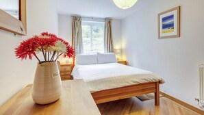 Une chambre avec un lit en bois, une table de chevet avec une lampe, un vase de fleurs, une fenêtre avec des rideaux et un tableau au mur.
