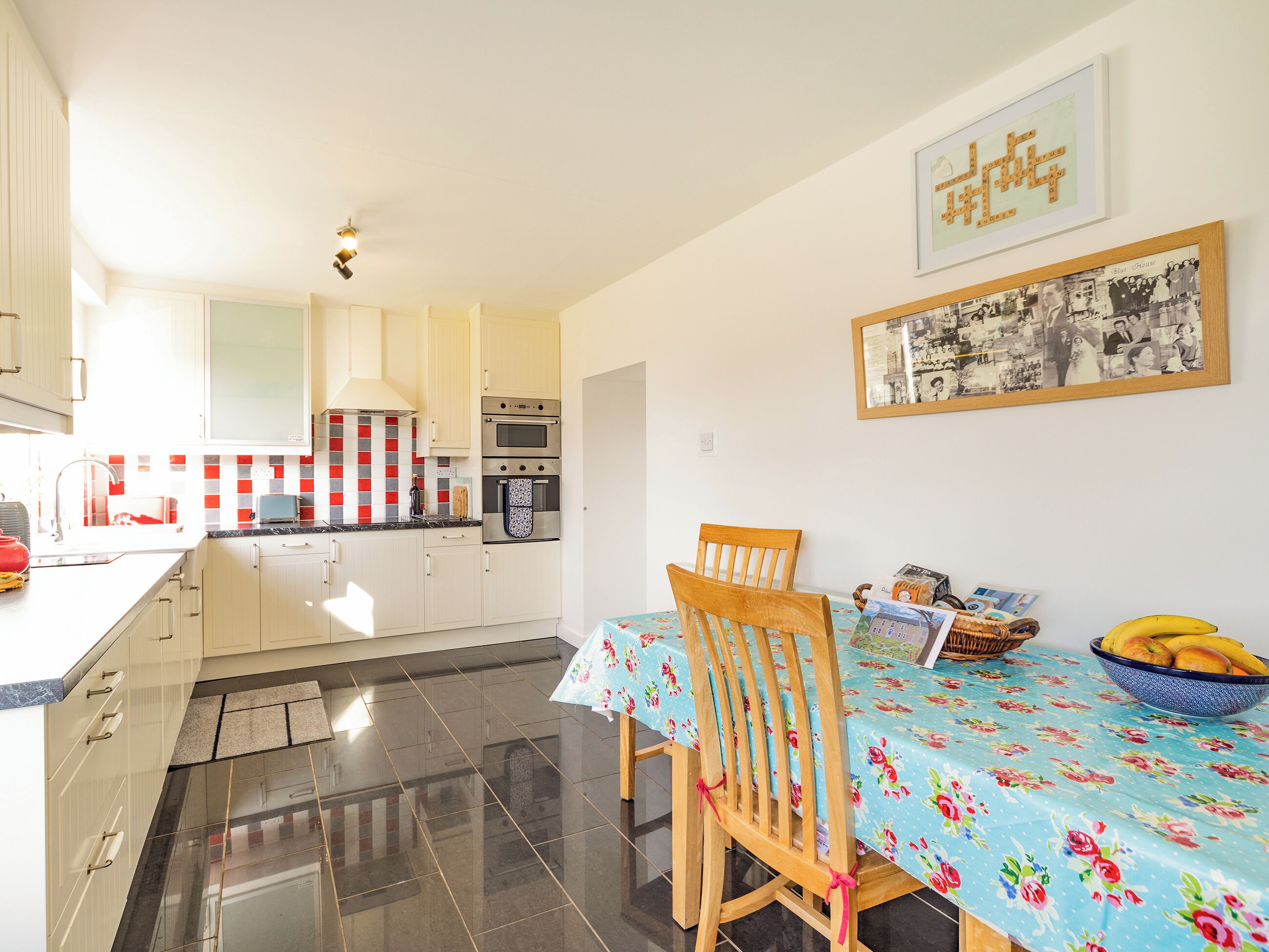 A modern kitchen with white cabinets, a red and white backsplash, and stainless steel appliances. There is a dining table with a floral tablecloth and a wooden chair. A framed picture hangs on the wall above the table.