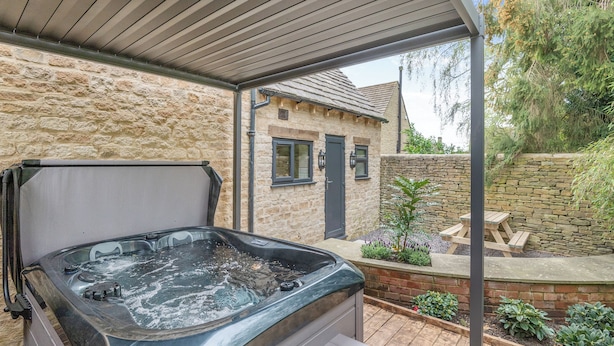 A hot tub with a wooden deck, stone walls, and a stone house in the background. - A luxury one bedroom cottage with bubbling hot tub. (South Cerney, near Cirencester)