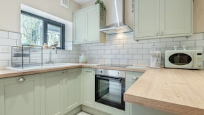A kitchen with light green cabinets, a stainless steel range hood, a microwave, and a window with a view of trees. - A luxury one bedroom cottage with bubbling hot tub. (South Cerney, near Cirencester)