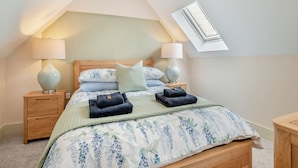 A bedroom with a sloped ceiling, a skylight, two bedside tables with lamps, a bed with a floral bedspread, and two folded towels. - A luxury one bedroom cottage with bubbling hot tub. (South Cerney, near Cirencester)