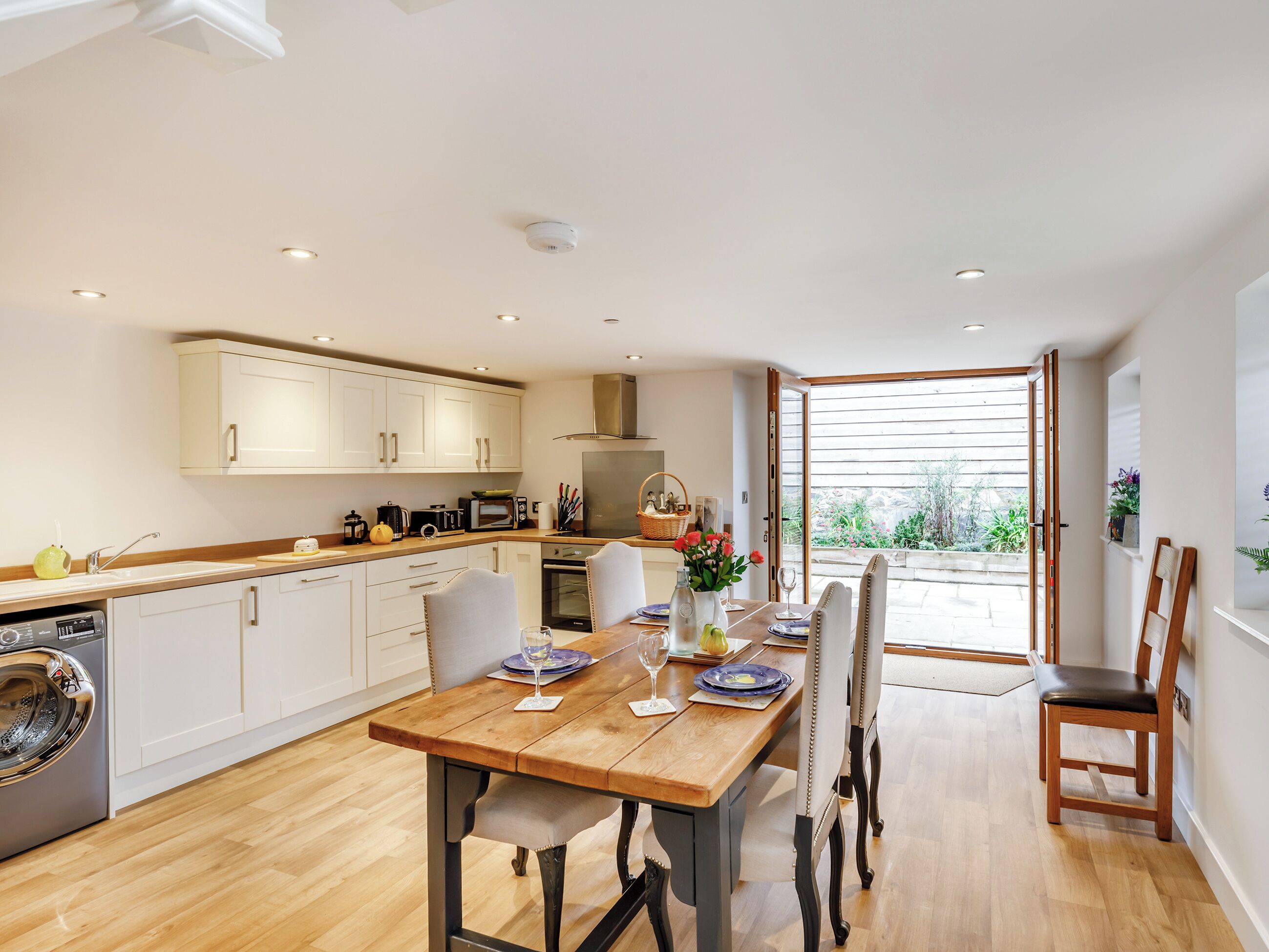 Una cocina moderna con armarios blancos, una mesa de comedor de madera y vista al jardín a través de una puerta abierta.