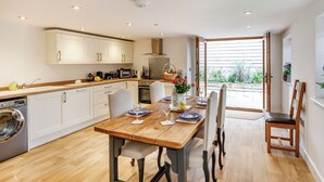 Una cucina moderna con mobili bianchi, un tavolo da pranzo in legno e vista sul giardino attraverso un'apertura.