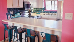 A modern kitchen with white cabinets, a stainless steel oven, a microwave, and a sink. There are three teal bar stools and a large island. - Pink Dream House in PCB - Beach, Pools, Hot Tub, Clubhouse, Dog-Friendly (Panama City)