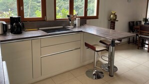 A kitchen with a central island, wooden cabinets, a coffee maker, and a view of greenery through large windows. - Small house on a hill, quiet with sea view, sunny terrace (Menton)