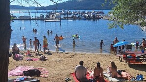 A lakeside beach with people swimming and sunbathing, a dock with boats, and a clear view of the lake and hills in the background. - House in Harrison with Lake View (Harrison)