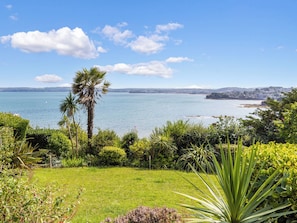 A coastal landscape with a grassy foreground, a palm tree, and a view of the sea and distant cityscape.