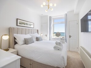 A neatly arranged bedroom with a bed, bedside tables, a framed picture, a mirror, and a view of the sea through a window.