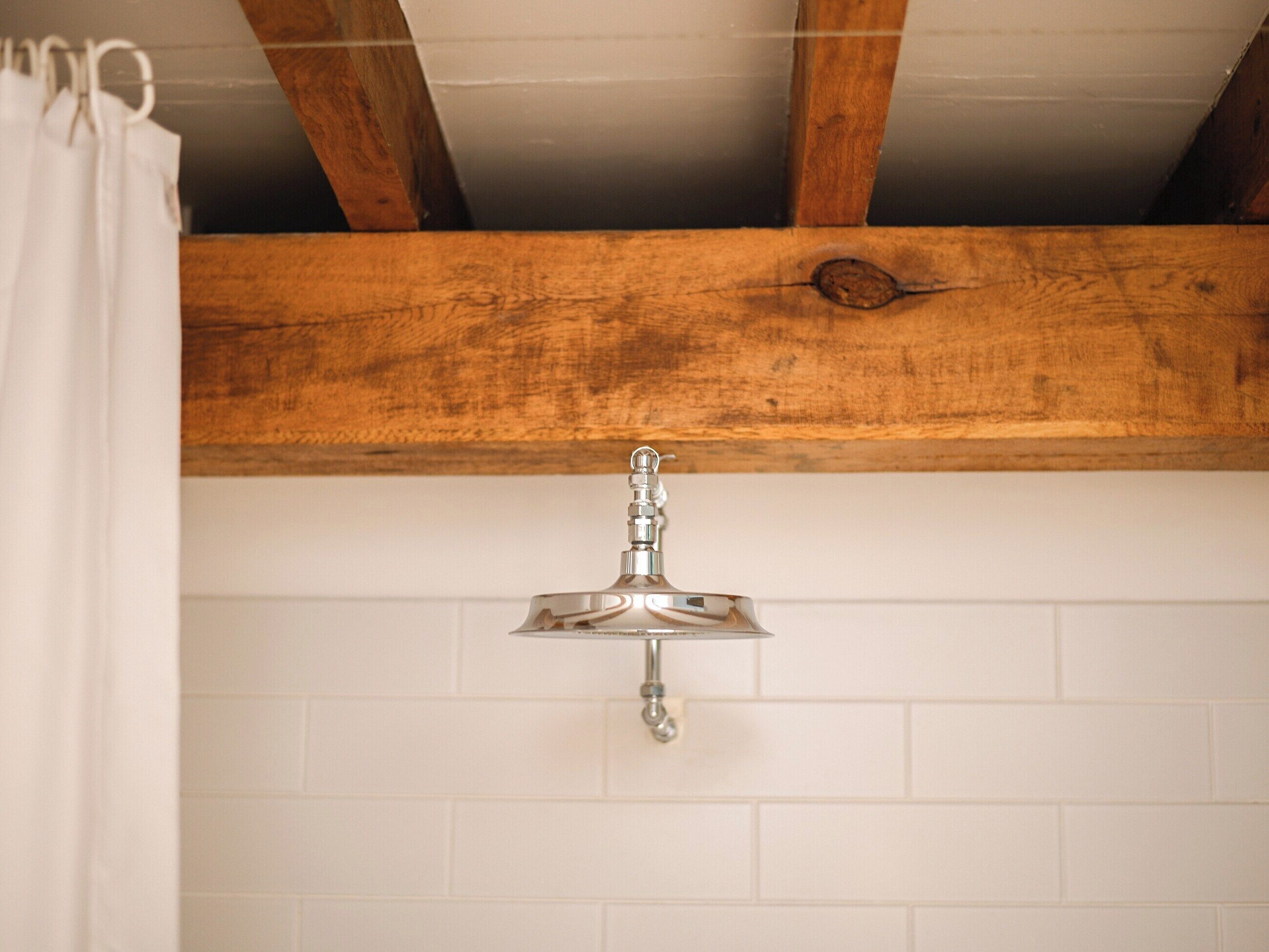 Een badkamer met een witte betegelde muur, een houten balk in het plafond en een zilveren kraan.