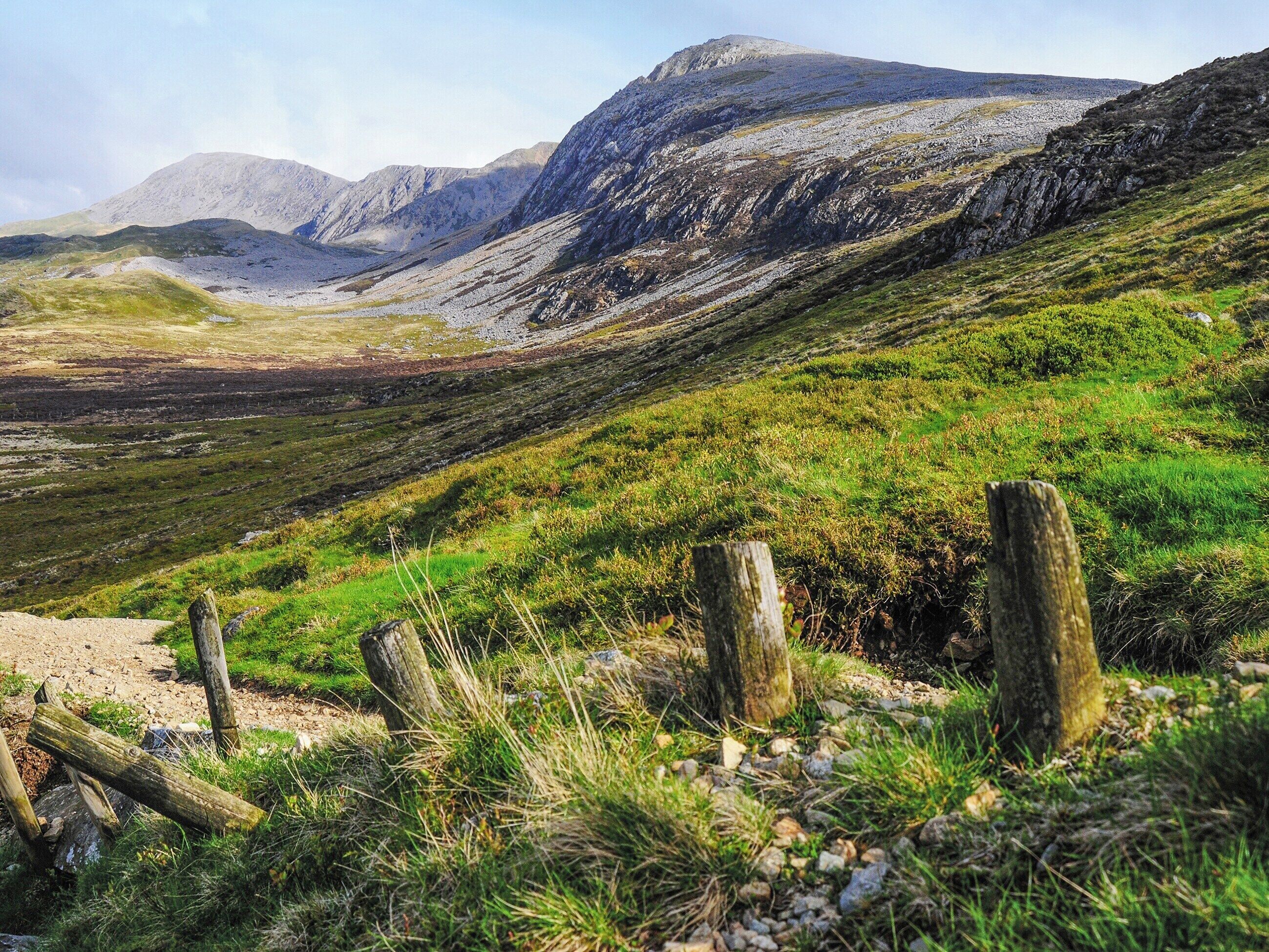 A mountainous landscape with a dirt path, wooden fence, and sparse vegetation.
