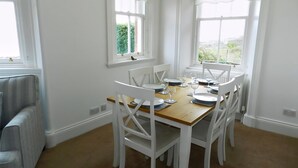 A dining area with a wooden table and white chairs, large windows with curtains, and a view of greenery outside. - 3 bedroom accommodation in Corrie, Isle of Arran (Corrie, Isle of Arran)