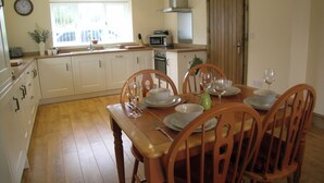 Een eetgedeelte met een houten tafel voor vier personen, witte kasten, een raam en een keuken op de achtergrond.