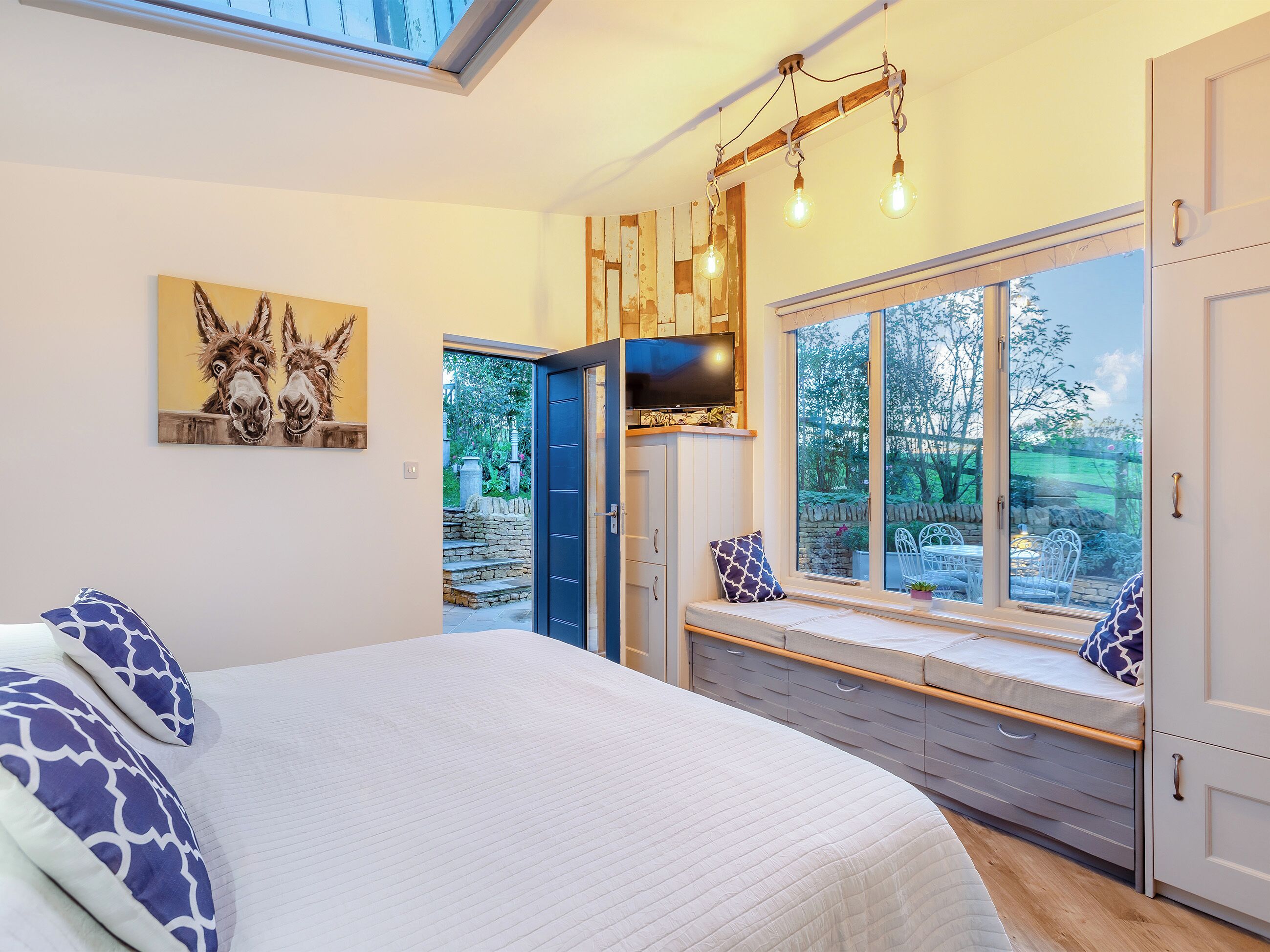 A bedroom with a bed, a bench with a TV, a large window, and a painting of donkeys.