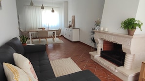 A modern living room with a dark sofa, a fireplace, and a dining area in the background. - Casa das Rosas- Villa with garden, Charneca da Cotovia (Charneca da Cotovia)