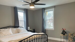 Un dormitorio con cama, ventilador de techo, dos ventanas con cortinas, una mesita con un jarrón de flores y una silla.