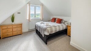 Un dormitorio con techo inclinado, una cama con colcha a cuadros grises y blancos, dos almohadas rojas, una mesita de noche con lámpara, un aparador de madera y una ventana con vistas a zonas verdes.