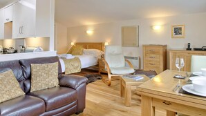 A modern hotel room with a leather sofa, a wooden dining table, a bed with white linens, a wooden cabinet, and a wall-mounted lamp.