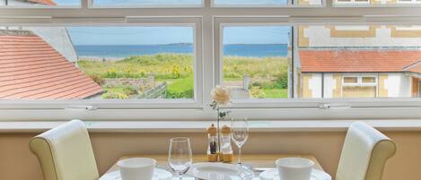 Une table à manger dressée pour deux personnes, avec vue sur la mer par la fenêtre.