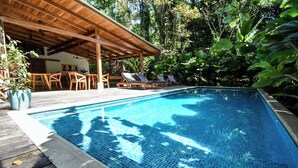 Um deck de madeira com piscina, espreguiçadeiras e área de estar coberta, tudo cercado por vegetação exuberante.