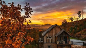 A rustic cabin with a balcony, surrounded by autumn foliage and a mountainous backdrop.