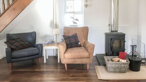 En hyggelig stue med en trætrappe, en sofa, en lænestol, et bord med bøger, en lampe, en pejs og en kurv med brænde.