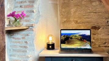 A laptop on a desk with a brick wall background, a wooden shelf with books, a vase with flowers, and a hanging light bulb.