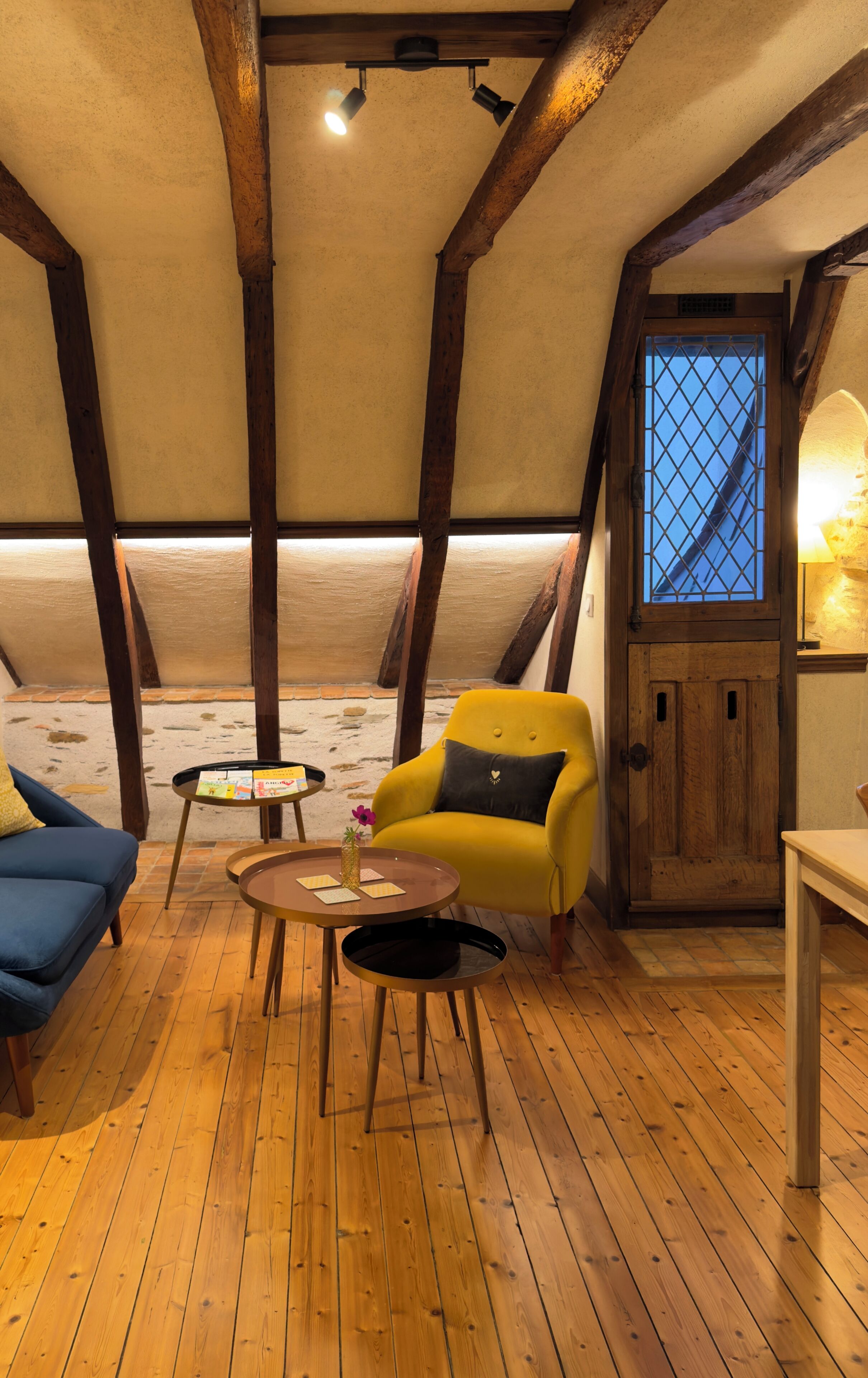 A cozy room with wooden beams, a yellow armchair, a small table with a vase, and a door with a window.