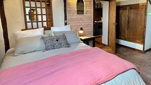 Une chambre à coucher avec un lit, des poutres en bois, un mur décoré d’un motif et une porte donnant sur la cuisine.