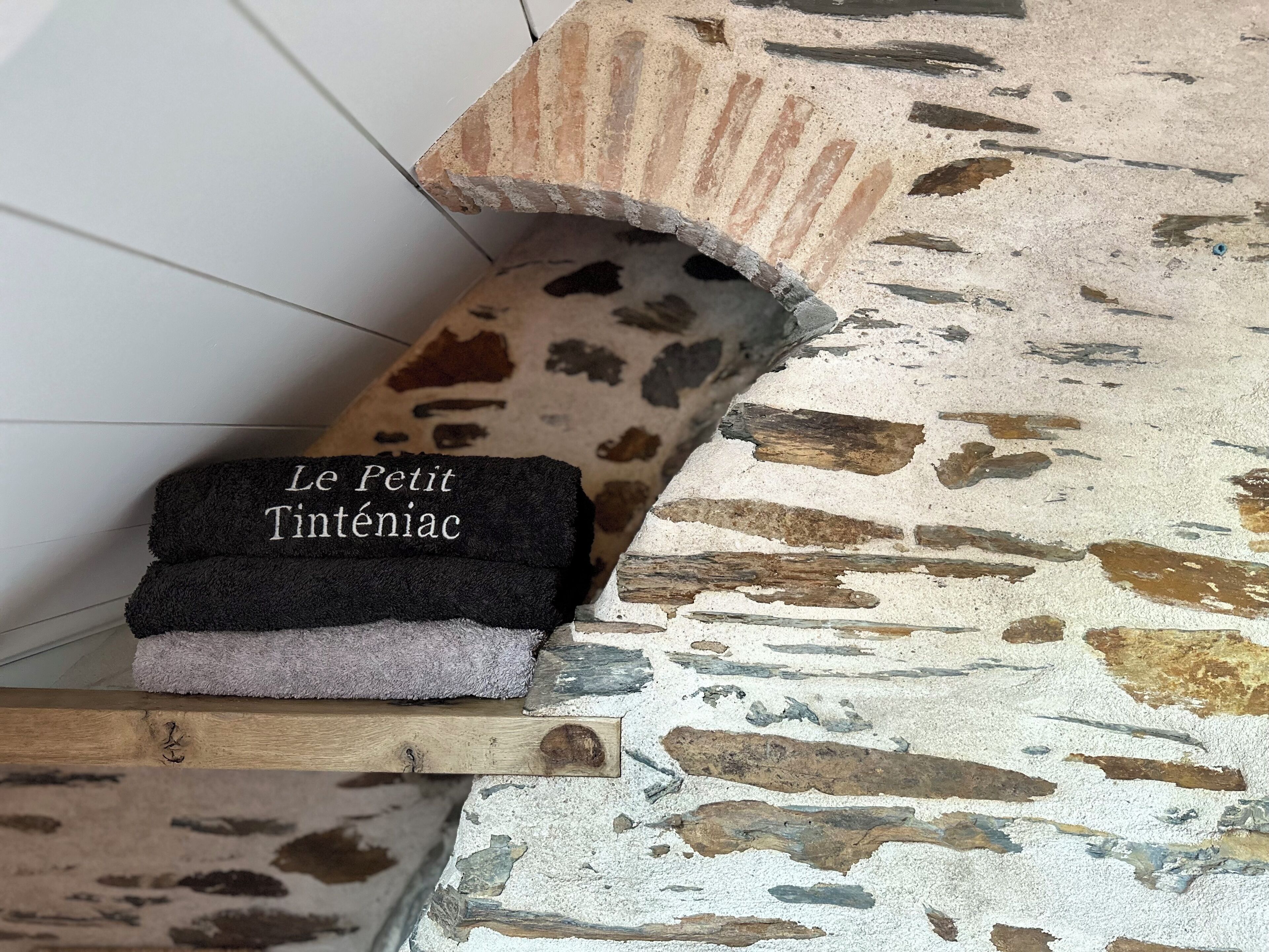 Ein Stapel Handtücher mit dem Schriftzug "Le Petit Inténiac" auf einem Regal vor einer strukturierten Wand.