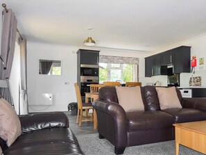 A living room with a brown leather sofa, a wooden dining table with chairs, and a kitchen area with dark cabinets.