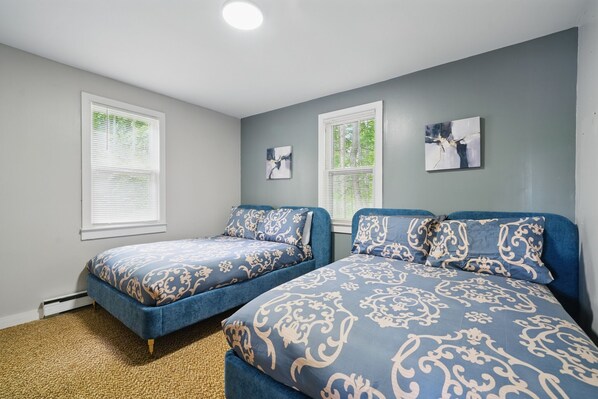A bedroom with two beds, each with a blue and white patterned bedspread, a small painting on the wall, and a window with blinds.