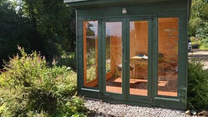 Une véranda en verre vert avec une terrasse en bois, entourée de verdure.