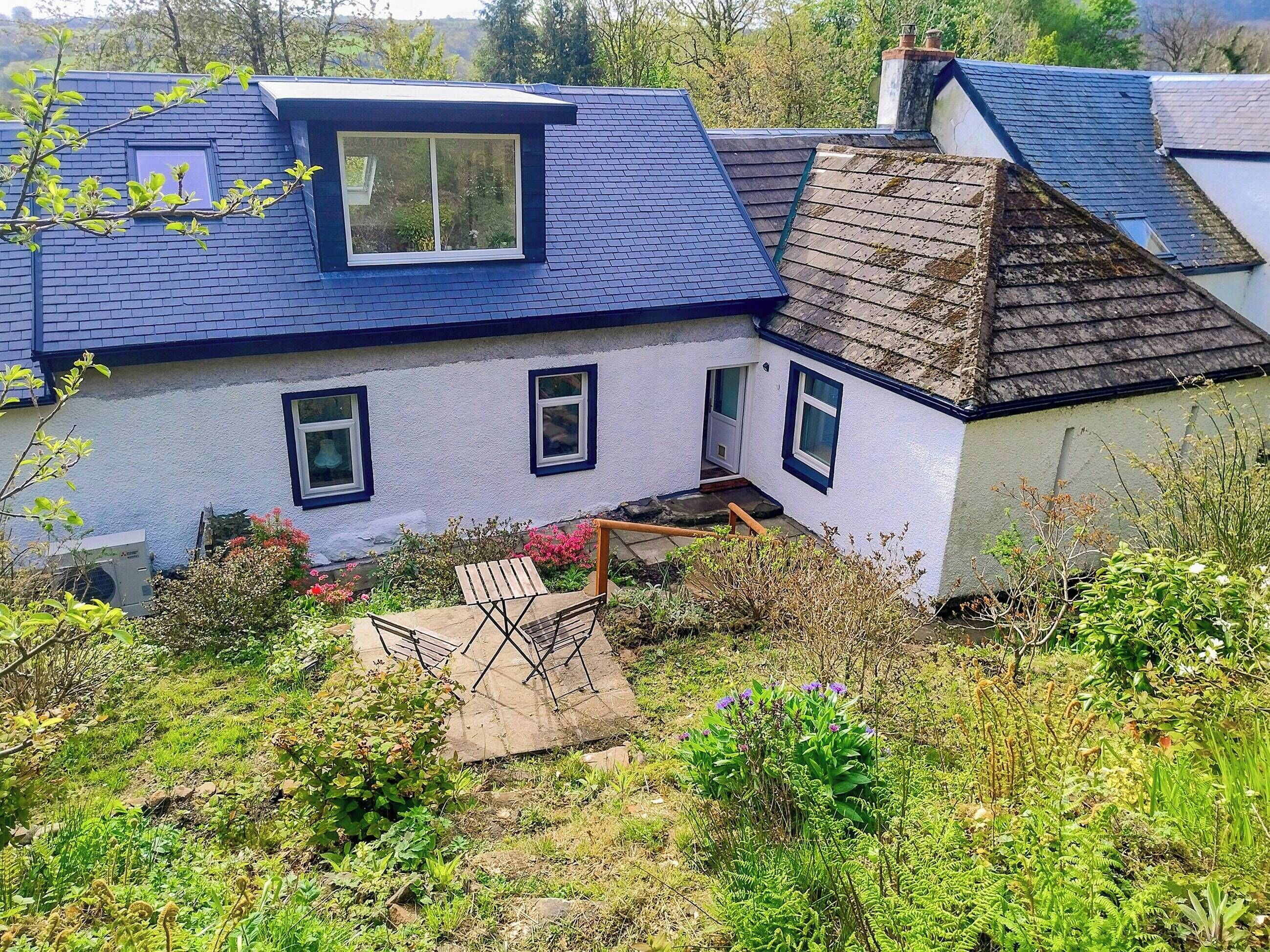 A traditional house with a sloped roof, a small garden with a wooden table and chairs, and a clear view of the surrounding greenery.